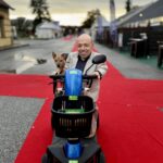 William Del Negro, presidente e fondatore di WIlleasy, mentre percorre un tappeto rosso a bordo di un mobility scooter blu, accompagnato da un piccolo cane seduto nel cestino anteriore. Sullo sfondo si intravedono le strutture di un evento all’aperto, con cielo nuvoloso e luce serale.