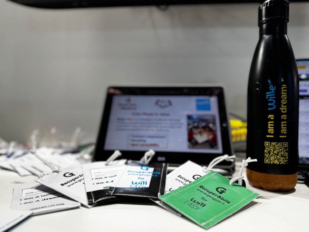Gadget Willeasy realizzati con materiali riciclati dal progetto RecuperAbile: cartellini in tessuto con messaggi e logo, accanto a una borraccia brandizzata, esposti durante un evento a Barcellona.