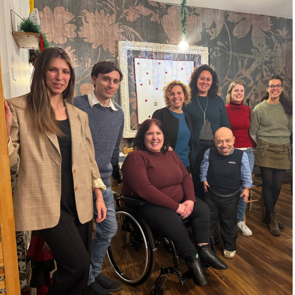 Nell&rsquo;immagine &egrave; ritratto un gruppo di persone durante un momento di team building di Willeasy, condotto da Umameta, in un ambiente caldo e accogliente: il Diversamente Bistrot, locale gestito da ANFFAS Udine. Il team &egrave; disposto in modo informale ma compatto, a sottolineare un forte senso di collaborazione e condivisione. I sorrisi e le posture rilassate dei partecipanti trasmettono un clima positivo, aperto e partecipativo. Al centro dell&rsquo;immagine emerge con chiarezza la diversit&agrave; del gruppo, sia in termini di genere sia di abilit&agrave;: la presenza di persone in piedi, sedute e di una persona in sedia a rotelle rappresenta concretamente i valori di inclusione, accessibilit&agrave; e valorizzazione delle differenze che sono alla base dell&rsquo;identit&agrave; di Willeasy. L&rsquo;abbigliamento &egrave; casual ma curato, coerente con un contesto lavorativo informale, mentre l&rsquo;arredamento e l&rsquo;illuminazione calda del Diversamente Bistrot contribuiscono a creare un&rsquo;atmosfera conviviale, ideale per favorire il dialogo, l&rsquo;ascolto e il rafforzamento delle relazioni. Nel complesso, la fotografia racconta un&rsquo;esperienza di team building autentica e significativa, in cui Umameta accompagna il team di Willeasy in un percorso di crescita condivisa, all&rsquo;interno di un luogo simbolico come il Diversamente Bistrot di ANFFAS Udine, che incarna concretamente i valori di inclusione sociale e partecipazione.