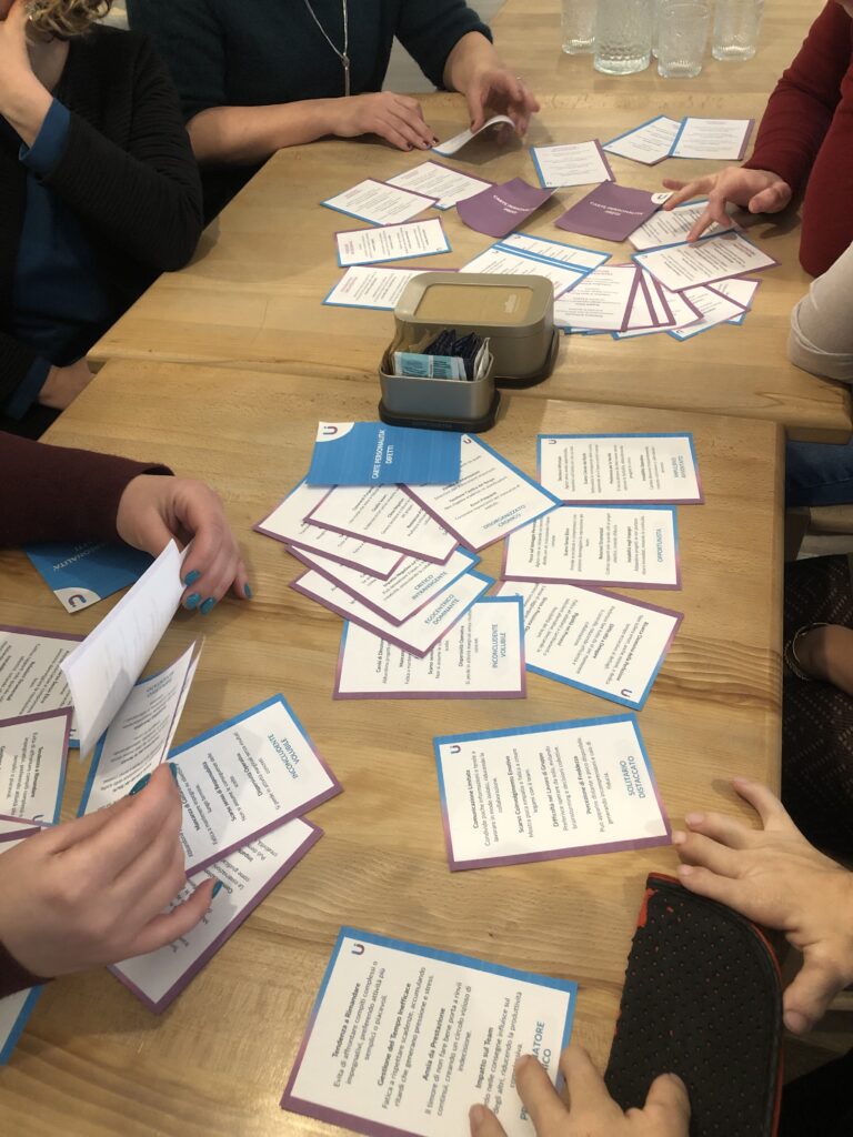 Nell&rsquo;immagine si vede un tavolo di legno con diverse mani intente a manipolare, leggere e sistemare numerose carte colorate. Le carte hanno bordi viola e azzurri e contengono testi stampati, probabilmente relativi a un&rsquo;attivit&agrave; o a un gioco strutturato legato a un percorso formativo. Al centro del tavolo c&rsquo;&egrave; anche una piccola scatolina metallica con bustine all&rsquo;interno. Il gruppo &egrave; concentrato e attivo, creando un&rsquo;impressione di collaborazione e scambio. L&rsquo;inquadratura &egrave; ravvicinata sulle mani e sui materiali, senza mostrare i volti, mettendo al centro l&rsquo;interazione e il lavoro condiviso.