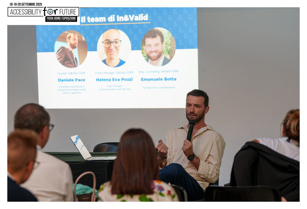 Fotografia scattata durante una presentazione all'evento "Accessibility For Future" presso la Fiera Udine Esposizioni. In primo piano, vista di schiena, si vede una parte del pubblico. Sullo sfondo, a destra, Daniele Pace, Founder di In&Valid, è seduto e parla a un microfono. Alla sua sinistra è proiettata una slide intitolata "Il team di In&Valid". La slide presenta tre foto con nomi e titoli: a sinistra, Daniele Pace (Founder In&Valid), al centro, Helena Eva Pozzi (Project Manager In&Valid), e a destra, Emanuele Botta (Resp. Counseling In&Valid).