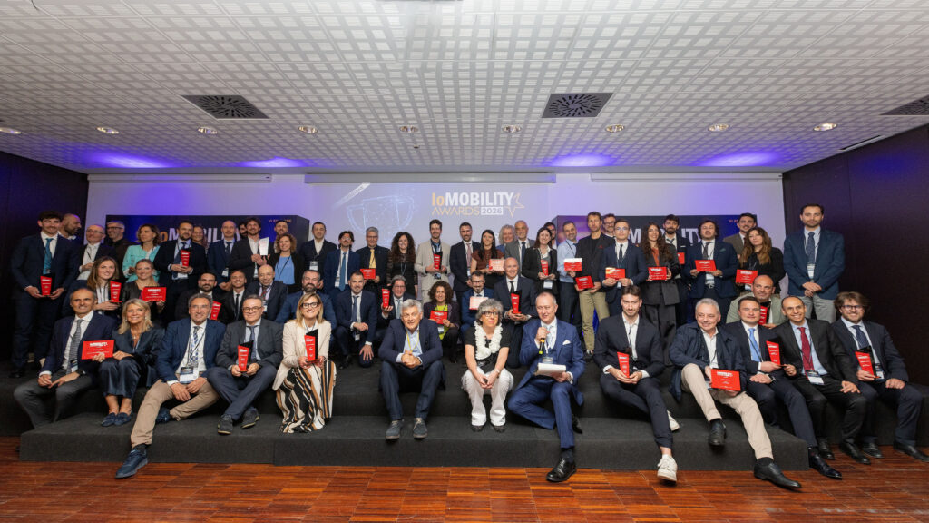 Foto di gruppo sul palco dei "ioMOBILITY AWARDS 2026". Numerosi premiati e relatori posano insieme tenendo in mano i loro trofei rossi. L'atmosfera è celebrativa e professionale.