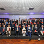 Foto di gruppo sul palco dei "ioMOBILITY AWARDS 2026". Numerosi premiati e relatori posano insieme tenendo in mano i loro trofei rossi. L'atmosfera è celebrativa e professionale.