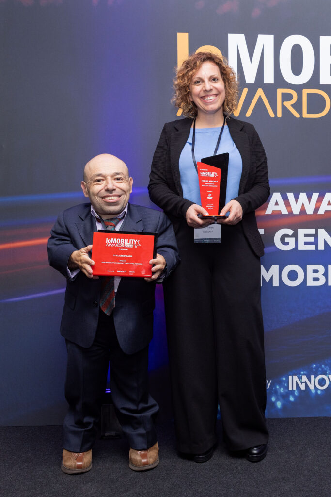 Primo piano di Gaia Citran e William Del Negro che tengono insieme un premio a forma di fiamma rossa ("Premio Speciale") davanti al logo dei "ioMOBILITY AWARDS 2026".