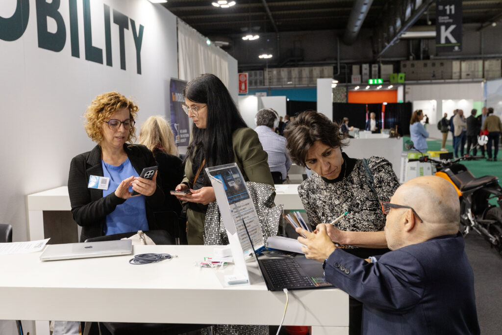 Un momento di lavoro e networking in una fiera. In primo piano, William Del Negro scrive su alcuni documenti sopra un tavolo bianco, mentre Gaia Citran e altre due persone accanto a lui controllano i propri smartphone o consultano materiali informativi.