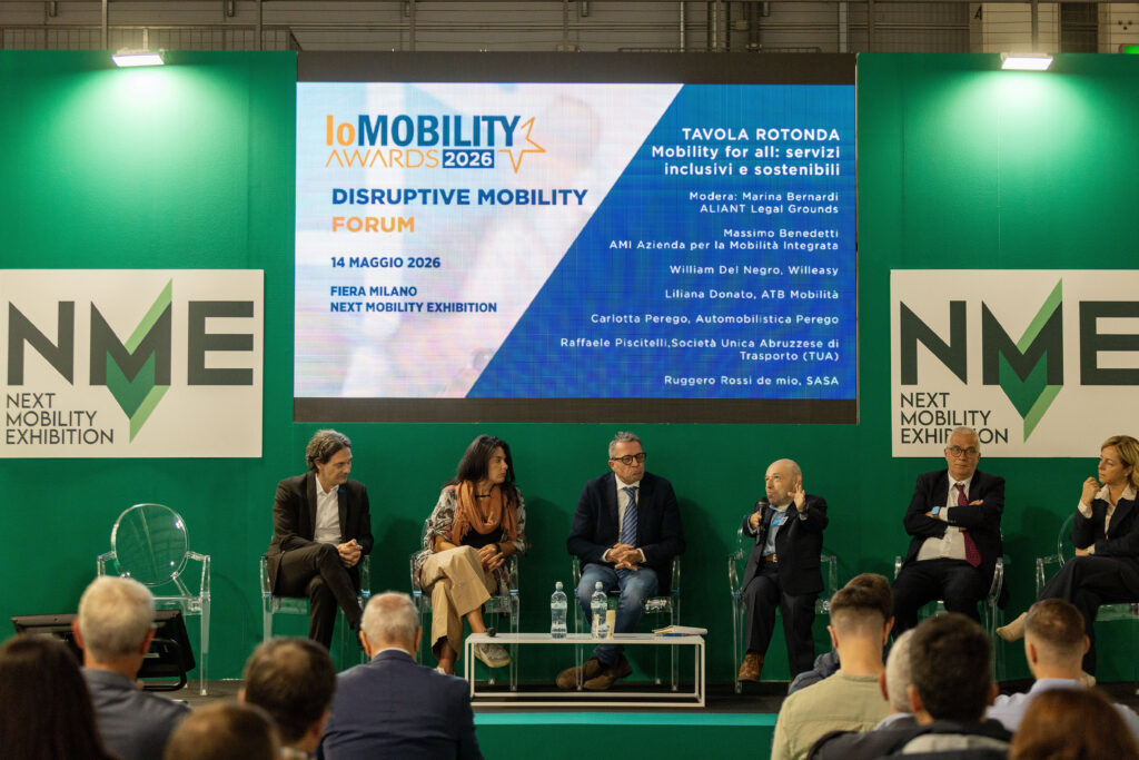 Una tavola rotonda intitolata "Mobility for all: servizi inclusivi e sostenibili". Sei relatori, tra cui William Del Negroche sta parlando al microfono, sono seduti su sedie trasparenti davanti a un grande schermo informativo al Next Mobility Exhibition.