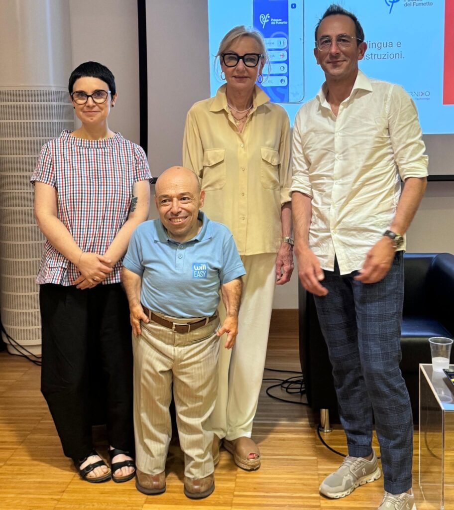 Ritratto di gruppo durante la presentazione di InclusivAPP. Da sinistra: Silvia Moras (Palazzo del Fumetto), William Del Negro (Willeasy), una relatrice ospite e Antonio Testa (AT Media). William Del Negro indossa una polo azzurra con il logo "will EASY". Sullo sfondo, uno schermo proietta una slide con il logo dell'app e la scritta "Palazzo del Fumetto - Lingua e Istruzioni".