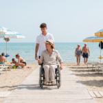 Uno stabilimento balneare italiano in una mattina di sole. Una passerella di legno chiaro attraversa la sabbia dall'ingresso fino al mare. Sulla passerella una donna di circa quarant'anni, con un cappello di paglia e un completo di lino chiaro, avanza sulla sua sedia a rotelle verso la riva. Sorride mentre parla con un giovane addetto dello stabilimento in polo bianca, che cammina accanto a lei. Ai lati ci sono file di ombrelloni a strisce azzurre e gialle e lettini bianchi. A sinistra una famiglia con bambini gioca sulla sabbia, a destra una coppia di persone anziane passeggia verso la battigia. Sullo sfondo il mare calmo e turchese.