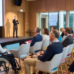 Sala conferenze luminosa con pareti in legno durante un evento aziendale. Sul palco un relatore con la giacca presenta davanti a un grande schermo con grafici colorati; accanto a lui un interprete della lingua dei segni, vestito di scuro, &egrave; ben visibile. In primo piano, a sinistra, una donna in sedia a rotelle segue l'intervento sorridendo. Il pubblico, composto da persone di et&agrave; diverse sedute su poltroncine grigie con dettagli azzurri, gialli e rosa, prende appunti su taccuini e tablet.