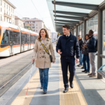 una donna cieca con il bastone cammina sulla striscia tattile di una fermata del tram, accanto a un addetto che parla con lei.